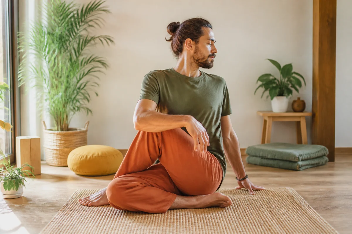 Adulte en torsion assise de tantra yoga dans une pièce lumineuse, entouré de plantes et d’accessoires de méditation